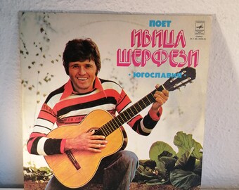 Puede incluir: Una portada de disco de vinilo con un hombre tocando la guitarra. La portada es rosa y blanca con el texto "POET IVICA ŠERFEZI - JUGOSLAVIJA" en negro.