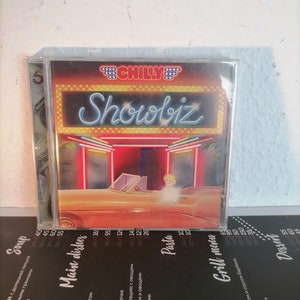 May include: A red and white neon sign with the text "Showbiz" in white letters. The sign is in front of a red and yellow building with a car parked in front. The car is yellow and has a red interior. The sign is in a clear plastic case.
