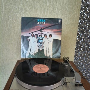 Op de afbeelding: Een vintage Lenco B55 platenspeler met een houten basis en een zwarte vinylplaat op het draaiplateau. Achter de platenspeler staat een ABBA vinylhoes met een foto van de bandleden die voor een helikopter staan bij zonsondergang.