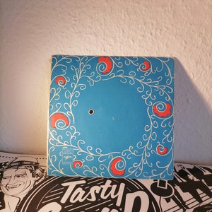 Puede incluir: Un disco de vinilo azul con un diseño floral blanco y rojo alrededor del borde. El centro del disco tiene un agujero para el eje.