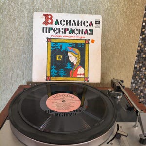 May include: A vintage vinyl record titled Vasilisa the Beautiful with a colorful illustration of a woman in a traditional headpiece, placed behind a black record on a Lenco of Switzerland turntable with a wooden base and silver control panel.