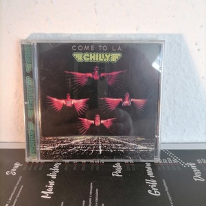Può includere: Una custodia per CD con sfondo nero e una band chiamata "CHILLY" in lettere bianche. Il nome della band è circondato da un design rosso, bianco e blu. La copertina dell'album presenta una città con quattro figure che volano nel cielo. Il titolo dell'album è "COME TO L.A."