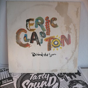 May include: A white vinyl record with a colorful painted design of the text "ERIC CLAPTON" and the text "Behind the Sun".