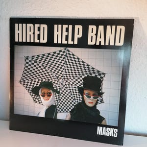 May include: A black and white vinyl record with the text "HIRED HELP BAND" and "MASKS" on the cover. The cover features a black and white checkered umbrella held by two mannequins wearing sunglasses and hats.