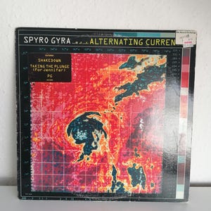 Puede incluir: Una portada de álbum de vinilo vintage de Spyro Gyra, "Alternating Currents". La portada presenta un gráfico vibrante de estilo mapa de calor en rojo, naranja y azul, con el nombre de la banda y el título del álbum en la parte superior. Hay una pegatina en la esquina superior derecha.