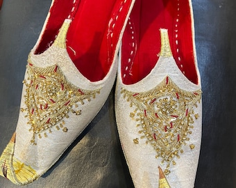 May include: A pair of white embroidered shoes with gold and red accents. The shoes have a pointed toe and a flat sole.