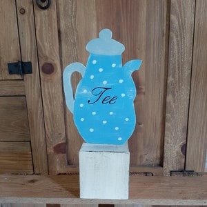 Könnte beinhalten: Eine blaue und weiße Teekanne mit weißen Punkten und dem Wort "Tee" darauf. Die Teekanne steht auf einem weißen Holzsockel.