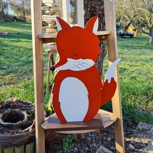 Peut inclure: Une figurine de renard en bois avec un design peint rouge et blanc. Le renard est debout sur une base en bois et a une expression d'œil fermé.