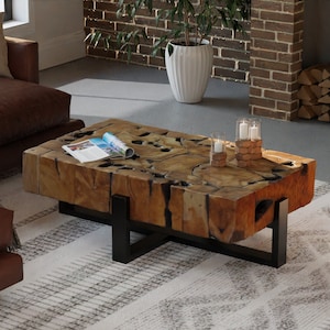 May include: A large, rustic coffee table made of reclaimed wood with a natural, brown finish. The table has a unique, organic shape and features a black metal frame. The table is sitting on a white and grey patterned rug in a living room setting.