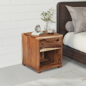 May include: A wooden bedside table with a drawer and open shelf. The table is made of natural wood with a warm brown tone. A small alarm clock, books, and a vase with greenery sit on top. The bedside table is next to a bed with pillows.