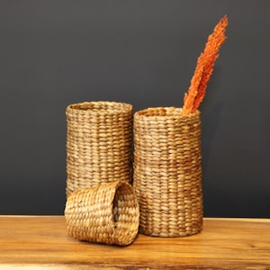Könnte beinhalten: Zwei geflochtene Körbe aus natürlichen Materialien, einer mit einem getrockneten Blumenstängel darin. Die Körbe sind zylindrisch und haben eine natürliche braune Farbe. Die Körbe stehen auf einer Holzoberfläche.