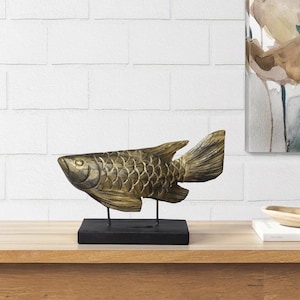 May include: A gold-coloured fish sculpture with detailed scales and fins, mounted on a black rectangular base. The sculpture is supported by two thin metal rods. The fish is displayed on a wooden surface, with a white brick wall in the background.