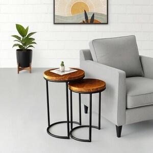 May include: Two round wooden side tables with black metal legs. The tables have a natural wood finish and are different sizes. A small plant and a magazine are on the larger table. The tables are in front of a gray armchair.