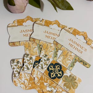 Peut inclure: Plusieurs étiquettes décoratives avec le texte "Jasmine's Mehndi" sont présentées. Les étiquettes ont un dessus crème avec un trou pour accrocher et un motif doré, vert et blanc. La partie inférieure des étiquettes présente un motif floral.