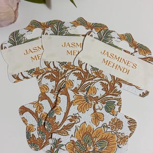 May include: Three decorative tags with a floral pattern in shades of yellow, brown, and green. Each tag has a cream-coloured label with the text "JASM ME" or "JASMINE'S MEHNDI" in a brown font. The tags are designed with a unique shape.