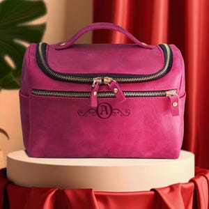 Puede incluir: Un neceser de viaje de color magenta con asa superior y cremalleras doradas. La bolsa tiene un ribete negro y una inicial decorativa "A" en la parte delantera. Se encuentra sobre un pedestal blanco con una tela de satén roja.