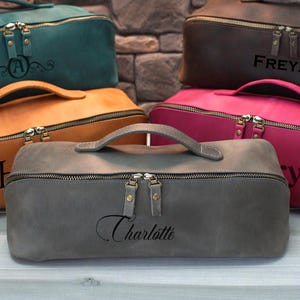 May include: A collection of personalized cosmetic bags in various colors, including teal, brown, pink, tan, and gray. Each bag features a zipper closure, a top handle, and custom text. The bags are arranged in a stacked display.