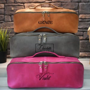 May include: Three rectangular zippered cosmetic bags in shades of brown, gray, and pink are stacked. The top bag is brown with "GRACE" printed on it. The middle bag is gray with "Charlotte" printed on it. The bottom bag is pink with "Violet" printed on it.