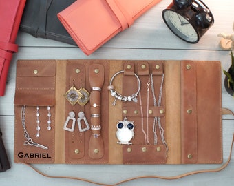 May include: A brown leather jewelry roll with multiple compartments for storing necklaces, earrings, bracelets, and rings. The roll is open and features a variety of jewelry, including a silver owl pendant necklace, a charm bracelet, and several pairs of earrings. The roll is labeled "GABRIEL".