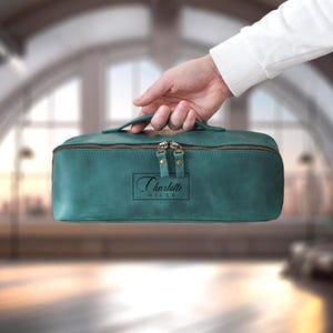 May include: Teal-colored leather travel bag with a top handle and a zipper closure. The bag has a rectangular shape and a small tag with the text "Charlotte Miller". The bag is held by a person.