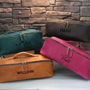May include: Four leather cosmetic bags in teal, brown, pink, and tan, each with a handle and a zipper closure. The bags are personalized with names: "Hazel", "Willow", and "Violet". The teal bag has initials.