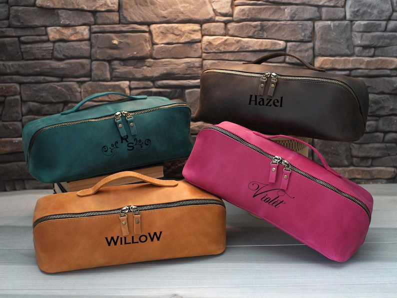 May include: Four leather cosmetic bags in teal, brown, pink, and tan, each with a handle and a zipper closure. The bags are personalized with names: "Hazel", "Willow", and "Violet". The teal bag has initials.