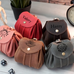 Pode incluir: Cinco sacos de dados de couro diferentes com nomes e designs personalizados. Os sacos s&atilde;o de v&aacute;rias cores, incluindo marrom, cinza, rosa e vermelho. Cada saco tem uma aba com fecho de press&atilde;o. Os sacos est&atilde;o sobre uma mesa com dados espalhados ao redor.