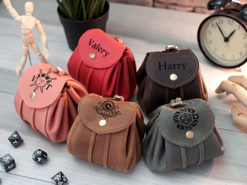 Pode incluir: Cinco sacos de dados de couro diferentes com nomes e designs personalizados. Os sacos s&atilde;o de v&aacute;rias cores, incluindo marrom, cinza, rosa e vermelho. Cada saco tem uma aba com fecho de press&atilde;o. Os sacos est&atilde;o sobre uma mesa com dados espalhados ao redor.