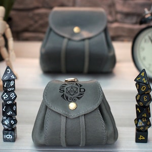 Pode incluir: Duas bolsas de dados de couro cinza com fecho de aba e bot&atilde;o dourado. Uma bolsa tem as letras "S D" e um s&iacute;mbolo de dado gravado na frente. Uma pilha de dados pretos com n&uacute;meros dourados e uma pilha de dados pretos e brancos com n&uacute;meros brancos est&atilde;o na frente das bolsas.
