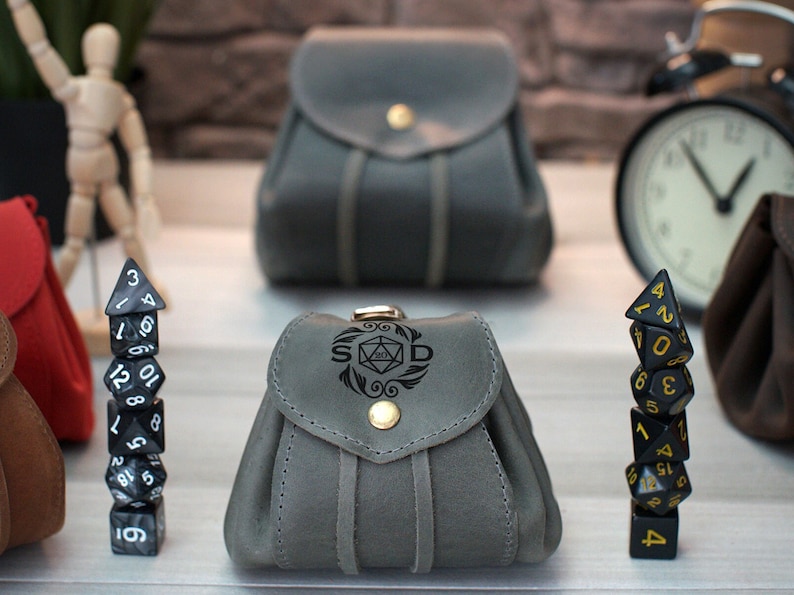 Pode incluir: Duas bolsas de dados de couro cinza com fecho de aba e bot&atilde;o dourado. Uma bolsa tem as letras "S D" e um s&iacute;mbolo de dado gravado na frente. Uma pilha de dados pretos com n&uacute;meros dourados e uma pilha de dados pretos e brancos com n&uacute;meros brancos est&atilde;o na frente das bolsas.