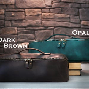 May include: Two leather travel bags, one dark brown and one teal, with top handles and zipper closures. The text "DARK BROWN" and "OPAL" are displayed above the bags. The bags are rectangular and sit on a surface in front of a brick wall.