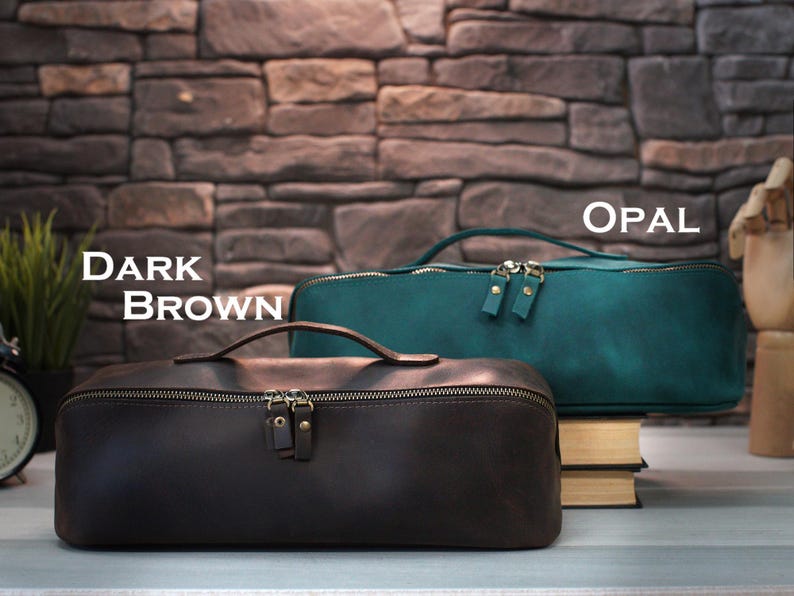 May include: Two leather travel bags, one dark brown and one teal, with top handles and zipper closures. The text "DARK BROWN" and "OPAL" are displayed above the bags. The bags are rectangular and sit on a surface in front of a brick wall.