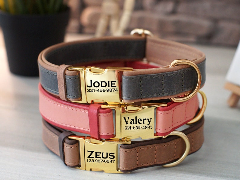 May include: Three leather dog collars stacked on top of each other. The top collar is gray with a tan trim, the middle collar is pink with a red trim, and the bottom collar is tan with a brown trim. Each collar has a gold buckle with a personalized name and phone number. The top collar says "JODIE 321-456-9874", the middle collar says "Valery 321-654-1895", and the bottom collar says "ZEUS 123-987-6547".