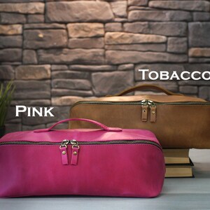 May include: Two leather travel bags, one pink and one tobacco brown, are displayed against a stone wall backdrop. The pink bag is in the foreground, with a handle and a zipper. The tobacco bag is behind it. The word "TOBACCO" is written above the brown bag.