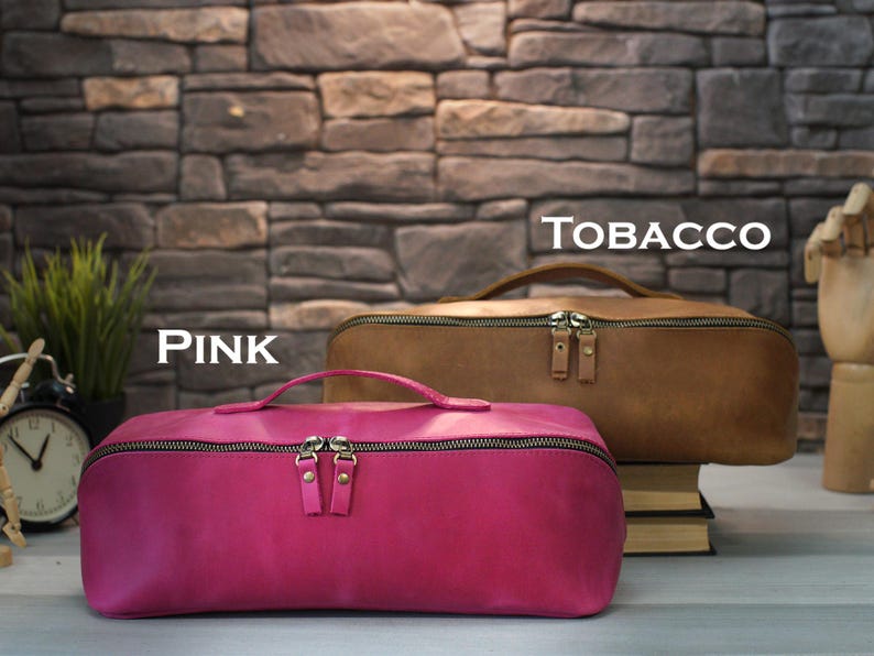 May include: Two leather travel bags, one pink and one tobacco brown, are displayed against a stone wall backdrop. The pink bag is in the foreground, with a handle and a zipper. The tobacco bag is behind it. The word "TOBACCO" is written above the brown bag.