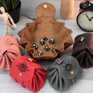 Pode incluir: Cinco sacos de dados de couro com nomes personalizados e uma bandeja de dados de couro marrom com dados pretos. Os sacos s&atilde;o rosa, vermelho, cinza e marrom.