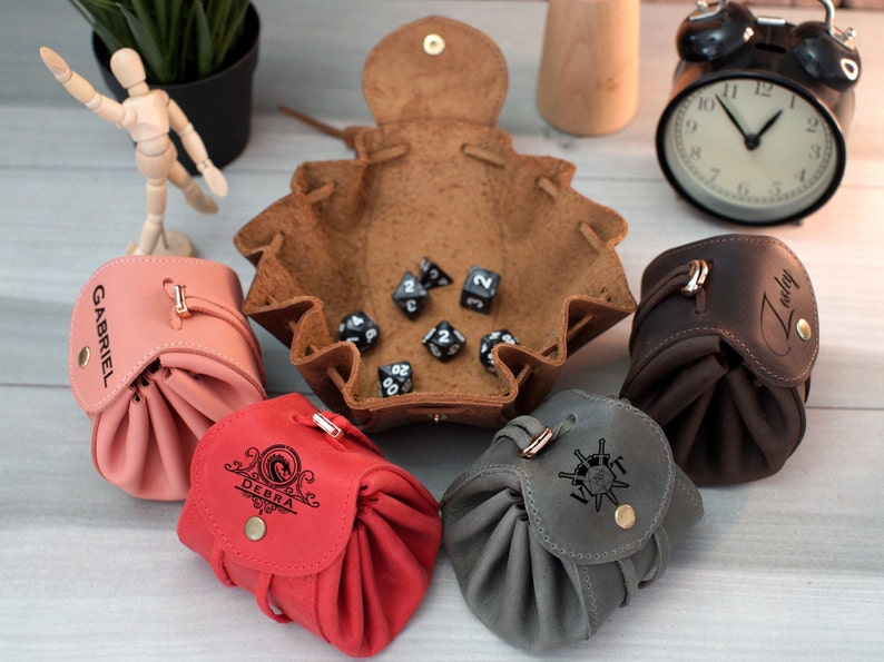 Pode incluir: Cinco sacos de dados de couro com nomes personalizados e uma bandeja de dados de couro marrom com dados pretos. Os sacos s&atilde;o rosa, vermelho, cinza e marrom.
