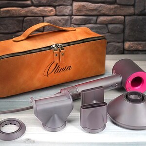 May include: A tan leather case with the name "Olivia" engraved on it, a gray and pink hair dryer, and several gray attachments are displayed on a light wood surface. The hair dryer has a pink accent.