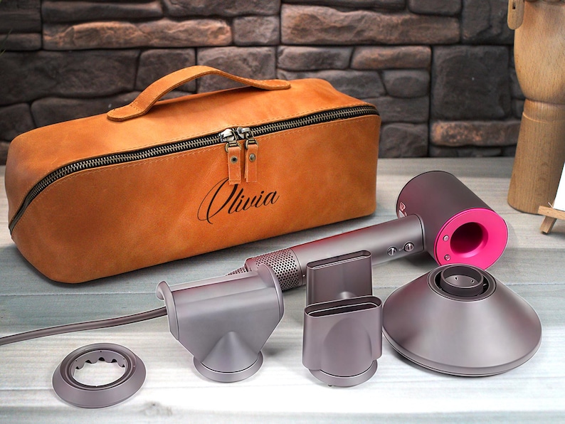 May include: A tan leather case with the name "Olivia" engraved on it, a gray and pink hair dryer, and several gray attachments are displayed on a light wood surface. The hair dryer has a pink accent.