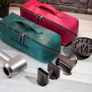 May include: A teal and a magenta leather travel case with a handle and zipper, with the name "FLORENCE" embossed on the teal case. A silver hair dryer and attachments are also visible, including a diffuser and nozzles.