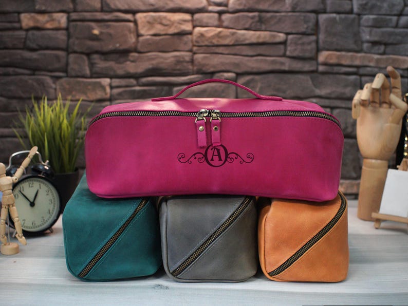 May include: A collection of leather cosmetic bags in various colors, including fuchsia, teal, gray, and orange. The fuchsia bag has a handle and is engraved with the letter "A" and decorative swirls. Each bag features a zipper closure.