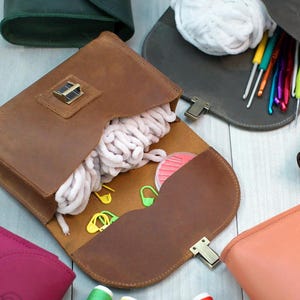 May include: A collection of brown leather bags with different colored interiors. The bags are open and show various items inside, including yarn, crochet hooks, and stitch markers.