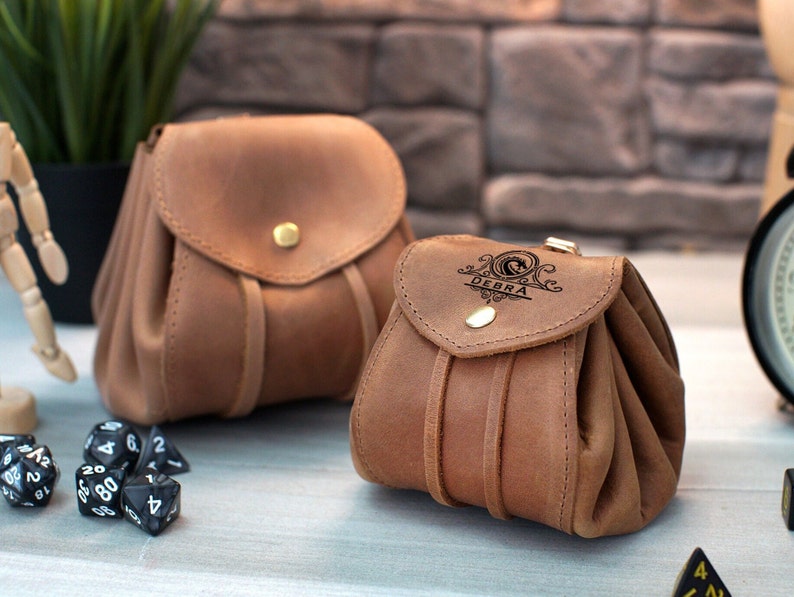 Pode incluir: Duas bolsas de couro marrom com cord&otilde;es. A bolsa maior &eacute; lisa, enquanto a bolsa menor tem o nome "DEBRA" gravado na aba frontal.
