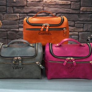 May include: Three leather cosmetic bags in orange, gray, and magenta. Each bag has a top handle, a wraparound zipper, and a name engraved on the front. The orange bag says "Gabriel", the gray bag says "Valery", and the magenta bag has initials.
