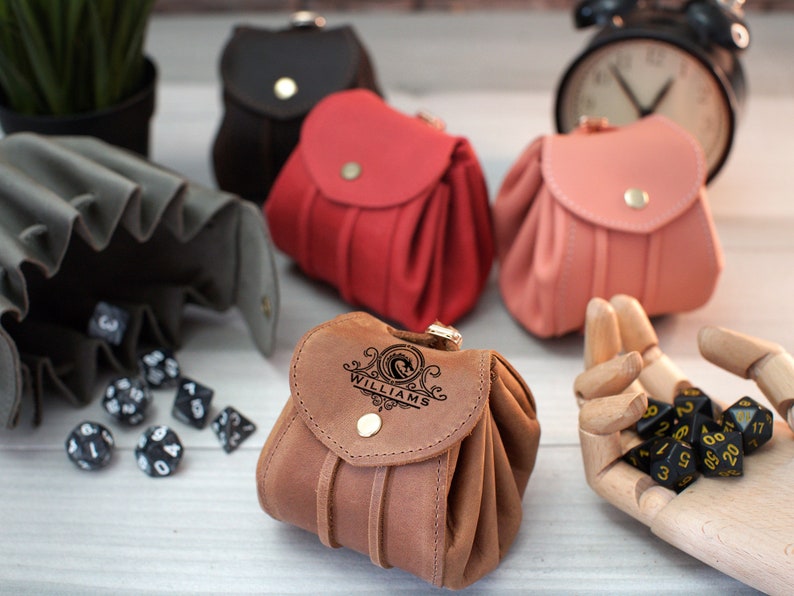 Pode incluir: Quatro sacos de dados de couro diferentes, um com o nome "WILLIAMS" gravado nele. Os sacos s&atilde;o marrom, vermelho, preto e rosa. H&aacute; tamb&eacute;m dados pretos em uma superf&iacute;cie de madeira.