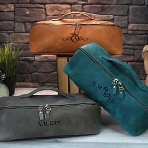 May include: Three leather travel bags in brown, teal, and gray, each with a top handle and a zipper closure. The brown bag has an "A" monogram, the teal bag has "RS", and the gray bag has "VALERY" engraved. The bags are on a wooden surface.