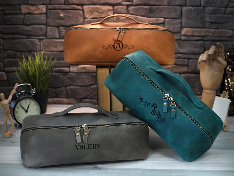 May include: Three leather travel bags in brown, teal, and gray, each with a top handle and a zipper closure. The brown bag has an "A" monogram, the teal bag has "RS", and the gray bag has "VALERY" engraved. The bags are on a wooden surface.