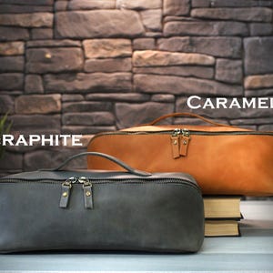 May include: Two leather pencil cases, one graphite gray and one caramel brown, are displayed. Each case has a zipper closure and a small handle. The graphite case is in the foreground, while the caramel case rests on a stack of books.