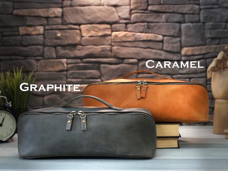 May include: Two leather pencil cases, one graphite gray and one caramel brown, are displayed. Each case has a zipper closure and a small handle. The graphite case is in the foreground, while the caramel case rests on a stack of books.