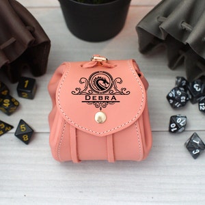 Pode incluir: Uma bolsa de dados de couro rosa com um design de drag&atilde;o e o nome "DEBRA" gravado na frente. A bolsa &eacute; cercada por dados pretos.